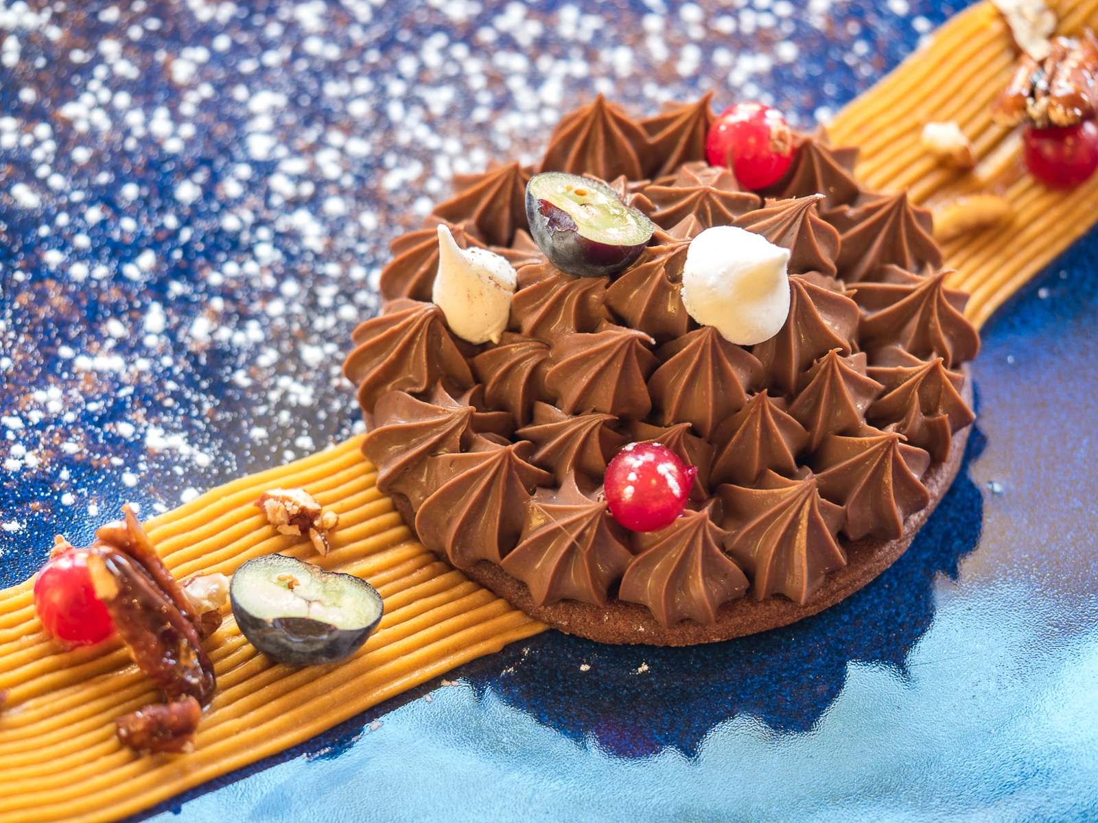 Dessert au chocolat garni de petites meringues, de raisins et de fruits, dressé dans une assiette bleue avec un élément jaune strié