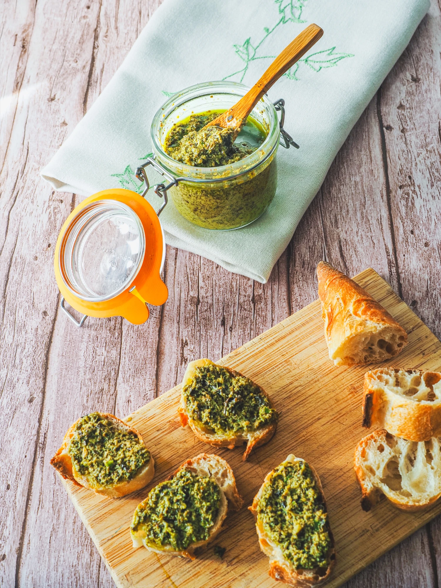 Bocal ouvert de pesto vert, tartines garnies et pain sur une planche en bois, photographiés du dessus
